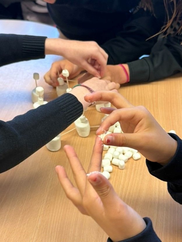 Four pairs of hands building small towers from marshmallows and toothpicks on a classroom table.
