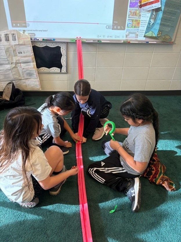 Four students kneeling on a classroom floor, assembling a long pink marble ramp.